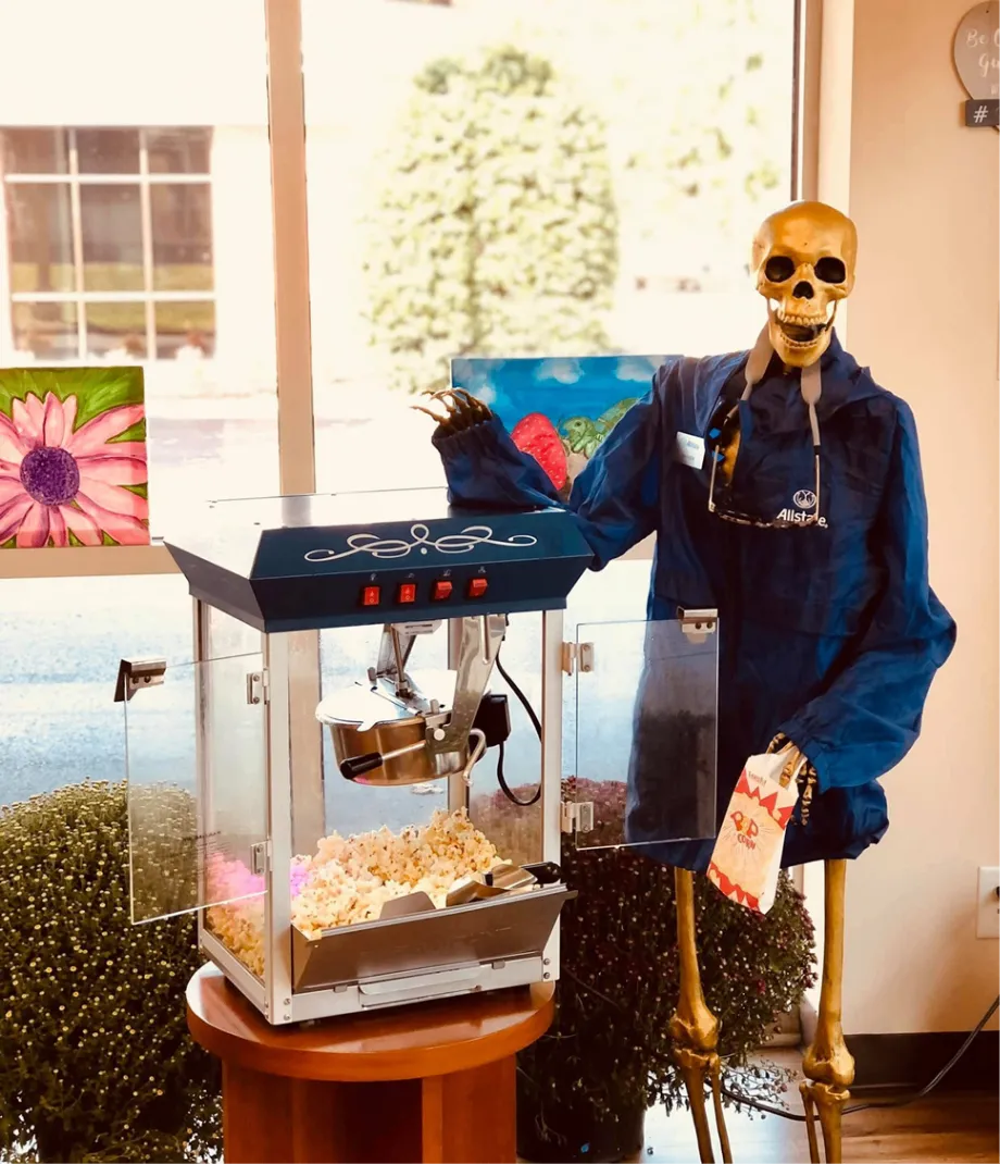 A skeleton in a blue jacket standing beside a popcorn machine and holding a striped popcorn bag