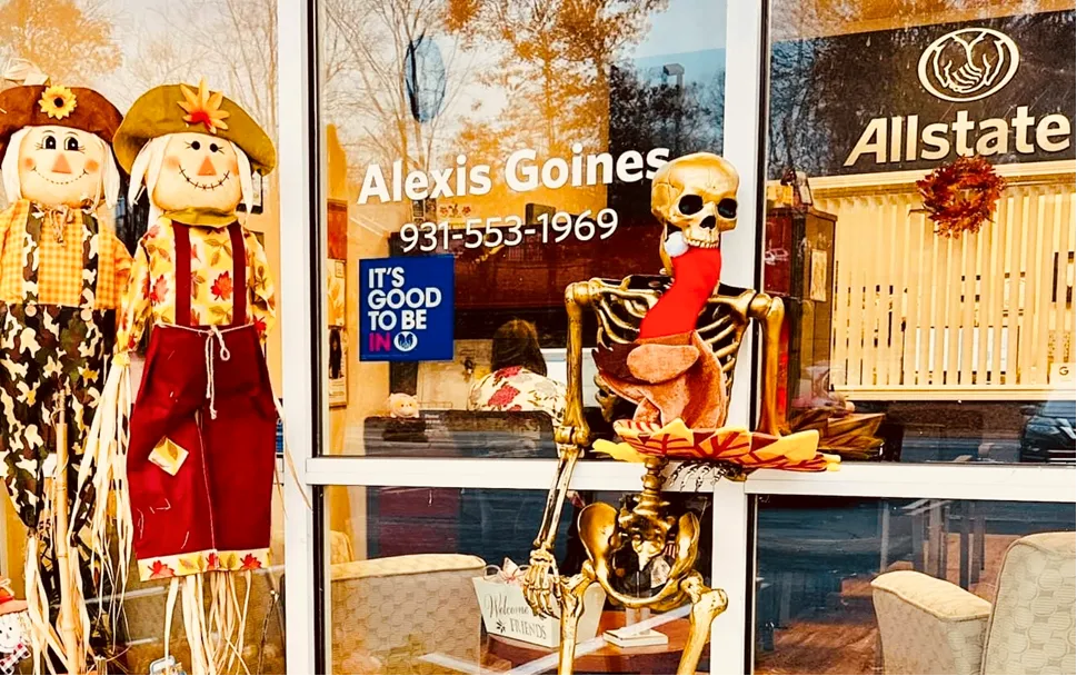 A seated skeleton in an office window display beside two scarecrows with an Allstate sign visible