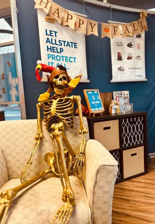 A skeleton in a colorful festive hat seated in a chair beneath a Happy Fall banner