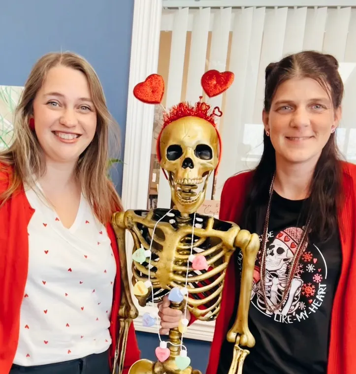 Two people standing beside a skeleton wearing a red heart headband and a string of heart decorations