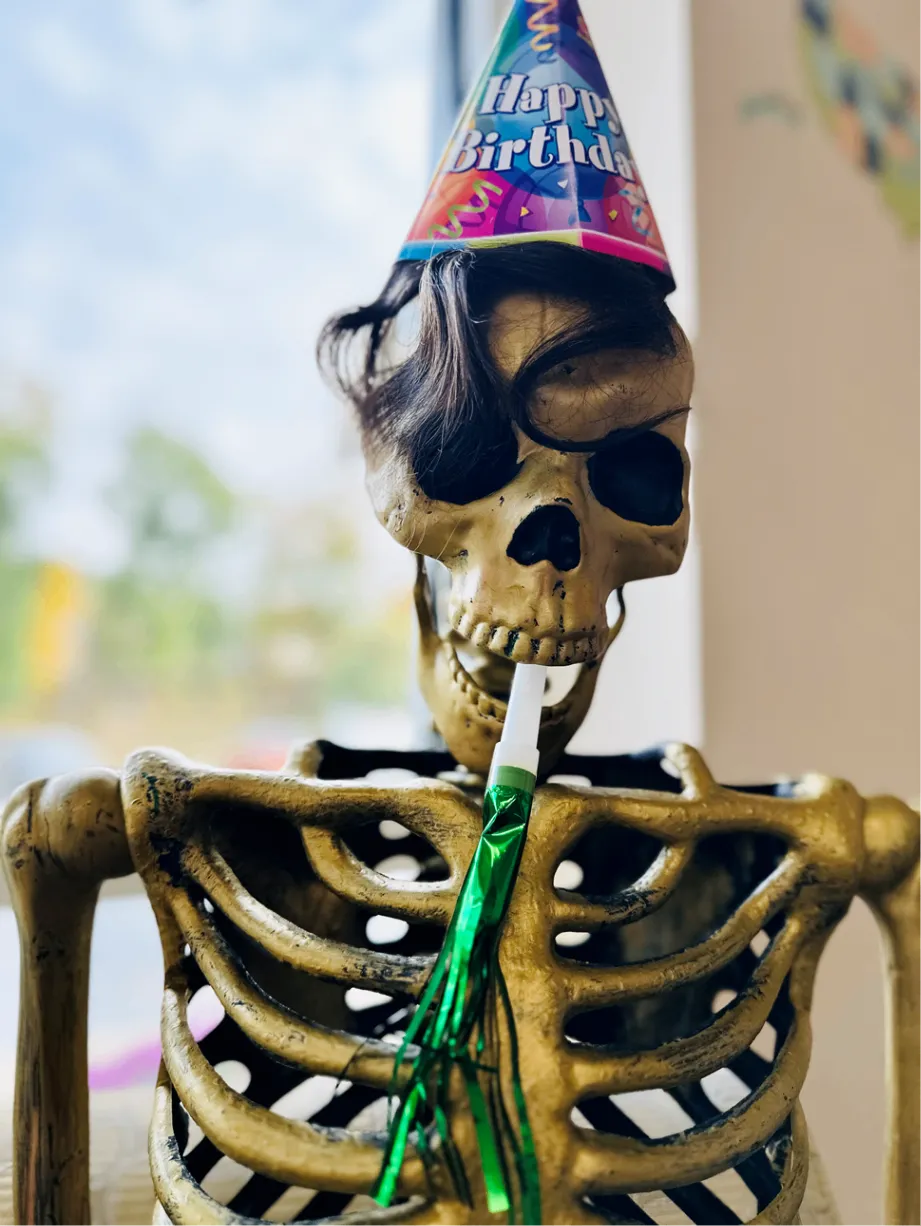 A close-up of a skeleton wearing a colorful Happy Birthday party hat and holding a party horn