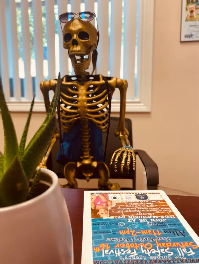 A skeleton wearing sunglasses seated behind an office desk with a plant and community flyer