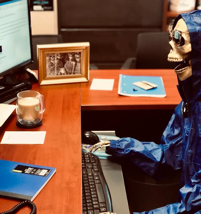 A skeleton in a blue jacket seated at an office desk beside a coffee cup and computer keyboard