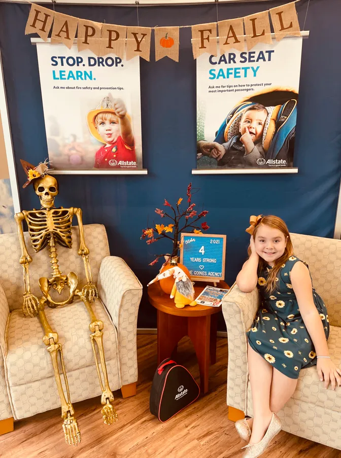 A skeleton and a child sit in separate armchairs beneath a Happy Fall banner and car-seat-safety posters in an office display