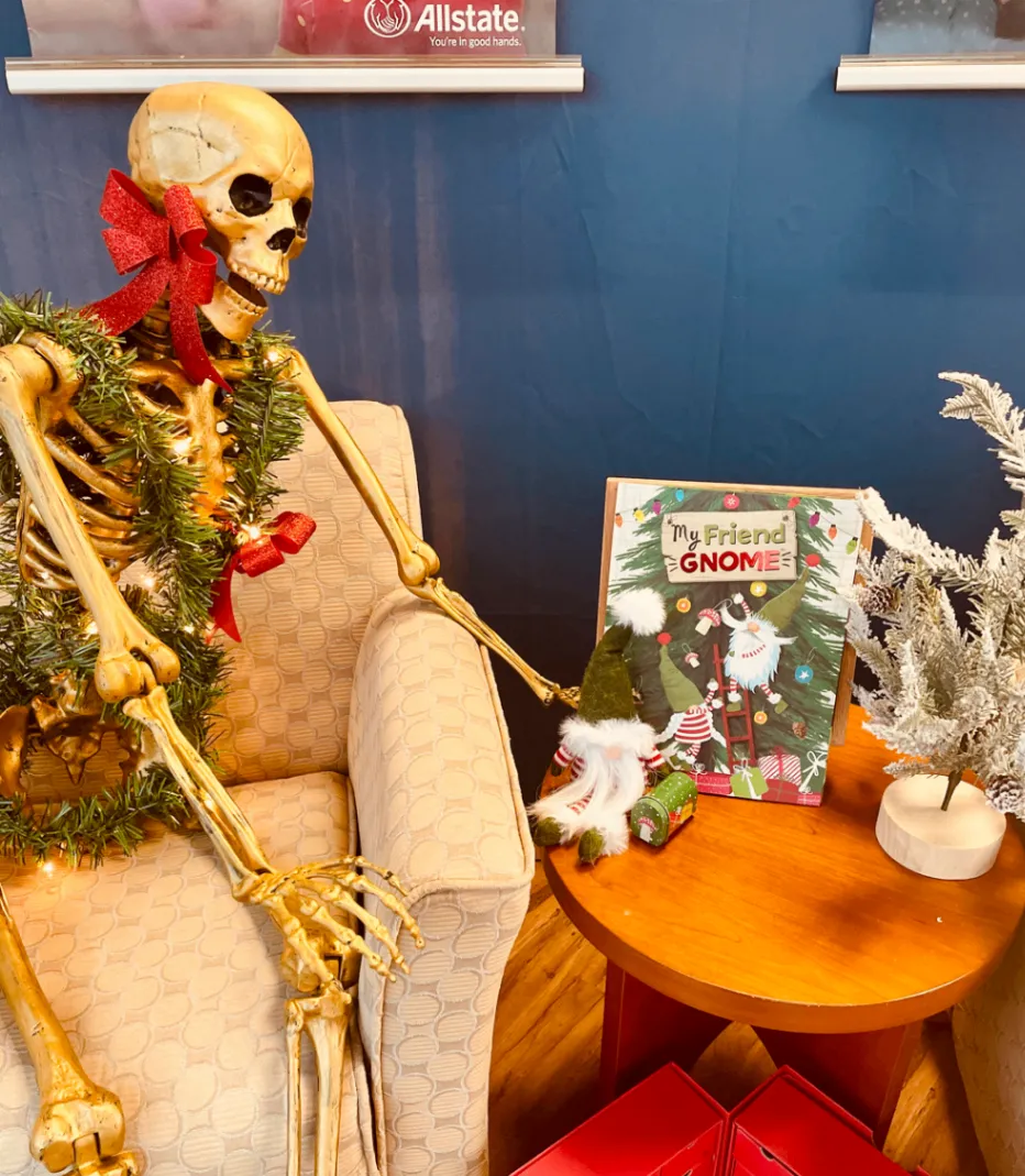 A skeleton wearing a red bow and green garland sits beside a holiday gnome display and a small white tree
