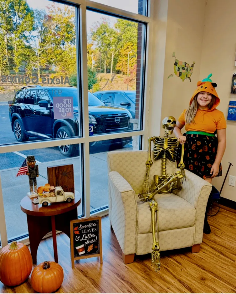 A seated skeleton in an armchair beside a young person wearing an orange pumpkin costume in a bright office window